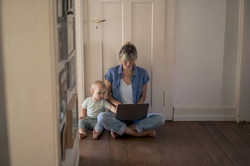 Working mother using laptop with son sitting on floor at home - JOSEF22417