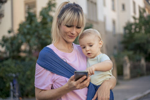 Son in baby sling using smart phone held by mother standing at street - JOSEF22419