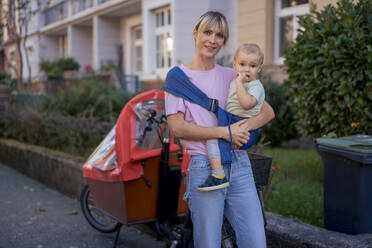 Smiling woman carrying son and standing near cargo bike - JOSEF22420