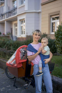 Woman carrying son in baby sling near cargo bike - JOSEF22421