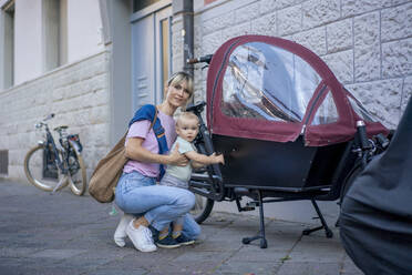 Mother crouching by son standing near cargo bike on sidewalk - JOSEF22422