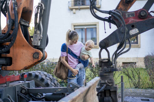 Mother carrying son standing near bulldozer - JOSEF22426