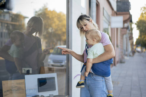 Mother pointing at store window with son in baby sling at street - JOSEF22427