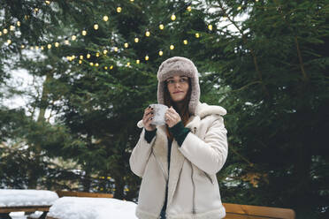 Frau in Winterjacke hält Kaffeetasse in der Hand in der Nähe von Bäumen - OLRF00003