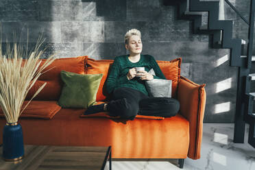Junge Frau mit Kaffeetasse, die zu Hause auf dem Sofa sitzt - OLRF00005