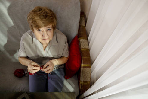Ältere Frau strickt auf einem Stuhl neben einem Vorhang zu Hause - MBLF00192