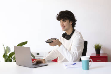 Smiling businessman talking on speaker phone and using laptop at home office - LMCF00707