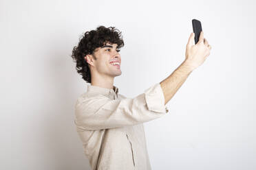 Smiling man taking selfie through smart phone against white background - LMCF00717