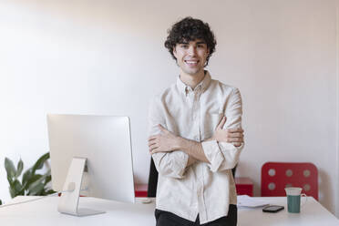 Smiling freelancer standing with arms crossed near desk at home office - LMCF00721