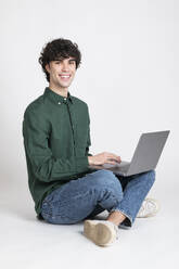 Lächelnder Mann sitzt mit Laptop vor weißem Hintergrund - LMCF00726