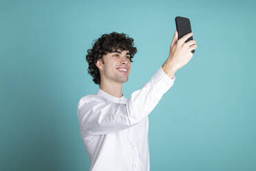 Lächelnder junger Mann nimmt Selfie durch Smartphone gegen cyan Hintergrund - LMCF00736