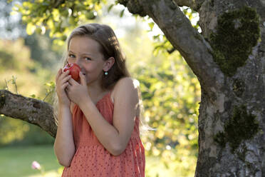 Smiling girl smelling apple near tree in garden - FLLF00901