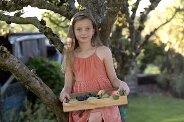 Lächelndes Mädchen hält Tablett mit Früchten in der Nähe von Baum im Garten - FLLF00902