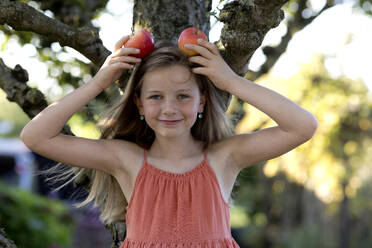 Lächelndes Mädchen hält Äpfel auf dem Kopf vor einem Baum - FLLF00904