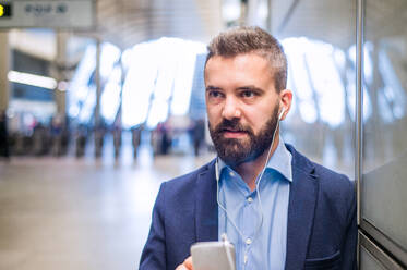 Close up of handsome hipster businessman with smartphone standing on subway station listening music - HPIF35890