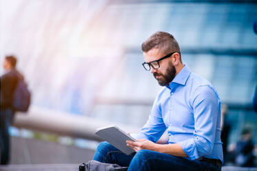 Hipster manager sitting on stairs on sunny day, working on tablet, London, City Hall - HPIF35912