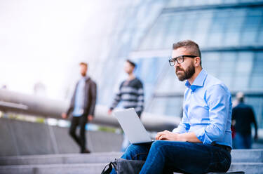 Hipster manager sitting on stairs on sunny day, working on laptop, London, City Hall - HPIF35913