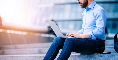 Unrecognizable hipster manager sitting on stairs on sunny day, working on laptop, London, City Hall - HPIF35915