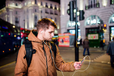Young hipster man with smart phone, listening music, walking in the streets of London at night - HPIF35923