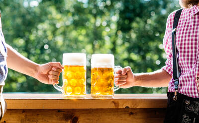 Zwei unerkennbare Männer in traditioneller bayerischer Kleidung halten Bierkrüge in der Hand und stoßen an. Oktoberfest. Sonniger Sommergarten. - HPIF35962