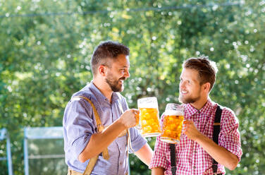 Zwei gut aussehende junge Männer in traditioneller bayerischer Kleidung halten Bierkrüge in der Hand und stoßen an. Oktoberfest. Sonniger Sommergarten. - HPIF35965