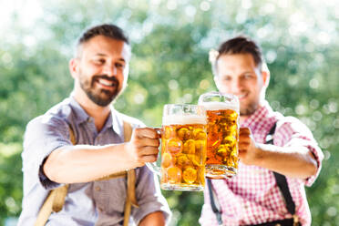 Zwei gut aussehende junge Männer in traditioneller bayerischer Kleidung halten Bierkrüge in der Hand und stoßen an. Oktoberfest. Sonniger Sommergarten. - HPIF35966