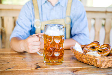 Unbekannter Mann in traditioneller bayerischer Kleidung sitzt am Tisch mit einem Krug Bier, Brezeln im Weidenkorb, Oktoberfest. - HPIF35971