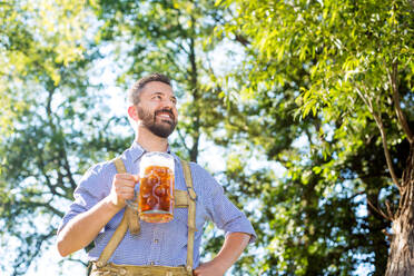 Hübscher junger Mann in traditioneller bayerischer Kleidung mit einem Bierkrug in der Hand, den Arm in die Hüfte gestemmt, Oktoberfest, sonniger Sommergarten. - HPIF35972