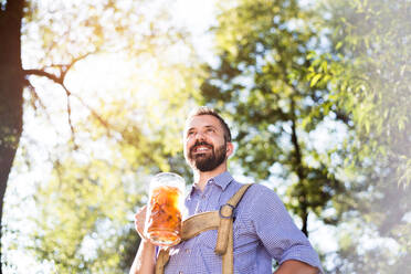 Hübscher junger Mann in traditioneller bayerischer Kleidung mit einem Bierkrug in der Hand. Oktoberfest. Sonniger Sommergarten. - HPIF35973