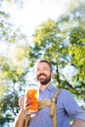 Handsome hipster young man in traditional bavarian clothes holding a mug of beer. Oktoberfest. Sunny summer garden. - HPIF35974