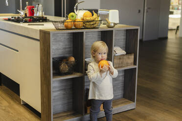 Girl holding orange fruit and standing near kitchen island at home - SEAF02110