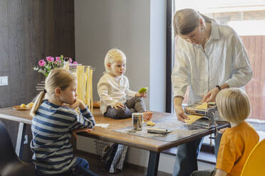 Woman cutting dough in pasta maker with family sitting at dining table - SEAF02123