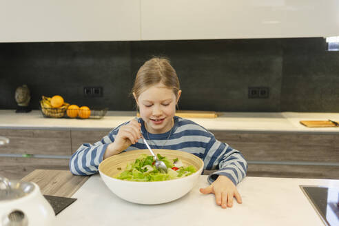 Lächelndes Mädchen isst Schüssel mit Salat mit Löffel in der Küche zu Hause - SEAF02136