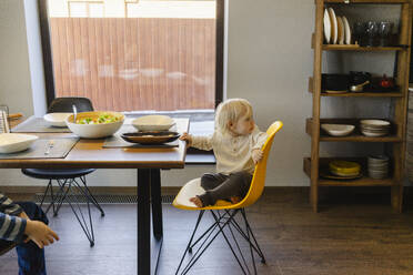 Blond girl sitting on yellow chair near dining table at home - SEAF02144