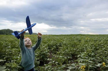 Smiling redhead boy playing with toy airplane in field - MBLF00200
