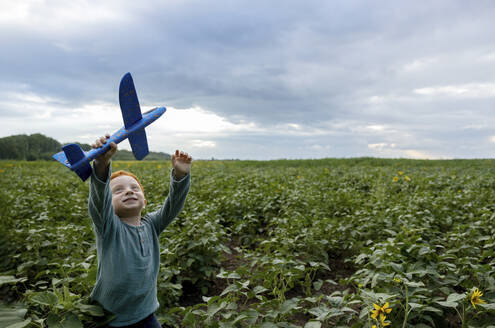 Lächelnder rothaariger Junge, der mit einem Spielzeugflugzeug auf einem Feld spielt - MBLF00200