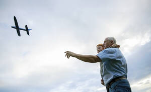 Lächelnder Großvater trägt Enkel und fliegendes Spielzeugflugzeug unter bewölktem Himmel - MBLF00203