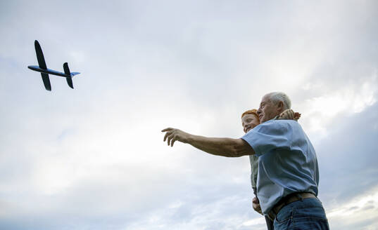 Smiling grandfather carrying grandson and flying toy airplane under cloudy sky - MBLF00203