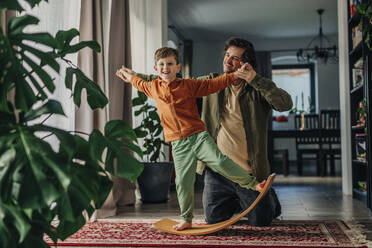 Smiling father helping son to balance on board at home - VSNF01530