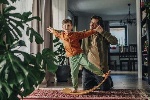 Smiling father helping son to balance on board at home - VSNF01530