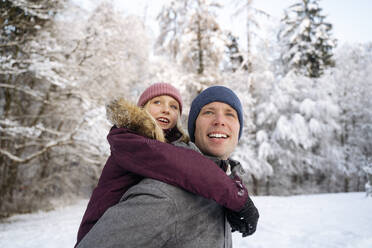 Lächelnder Mann, der seine Tochter im Winterwald huckepack nimmt - NJAF00723