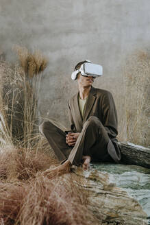 Geschäftsmann trägt Virtual-Reality-Simulator und sitzt auf einem Felsen in der Nähe einer Mauer im Garten - YTF01517