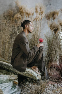 Nachdenklicher Geschäftsmann hält Nadelkissenblume und sitzt auf einem Felsen im Garten - YTF01521