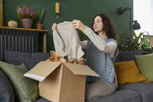Frau beim Auspacken eines Kleides aus einem Karton auf dem Sofa zu Hause - SECF00054
