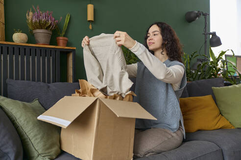 Woman unpacking dress from box sitting on sofa at home - SECF00054