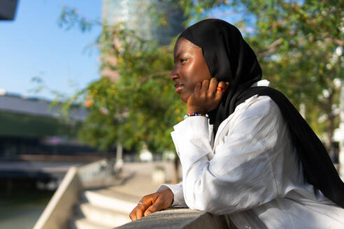 Seitenansicht einer nachdenklichen afroamerikanischen muslimischen Geschäftsfrau in schwarzem Hidschab, die ihr Kinn berührt und wegschaut, während sie sich auf ein Geländer in der Stadt stützt - ADSF50456