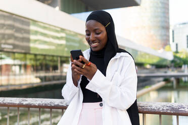 Cheerful African American young businesswoman in hijab browsing on smart phone while standing against modern building - ADSF50458