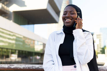 Cheerful African American young businesswoman in hijab with eyes closed talking over smart phone while standing against modern building - ADSF50459