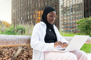 Seitenansicht einer ernsthaften, konzentrierten muslimischen Unternehmerin mit traditionellem Kopftuch, die auf einer Bank sitzt und einen Laptop benutzt, während sie an einem neuen Geschäftsprojekt arbeitet - ADSF50462
