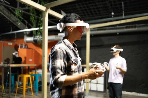 Side view of young excited man is engaged in a virtual reality game wearing a VR headset and using controllers - ADSF50475